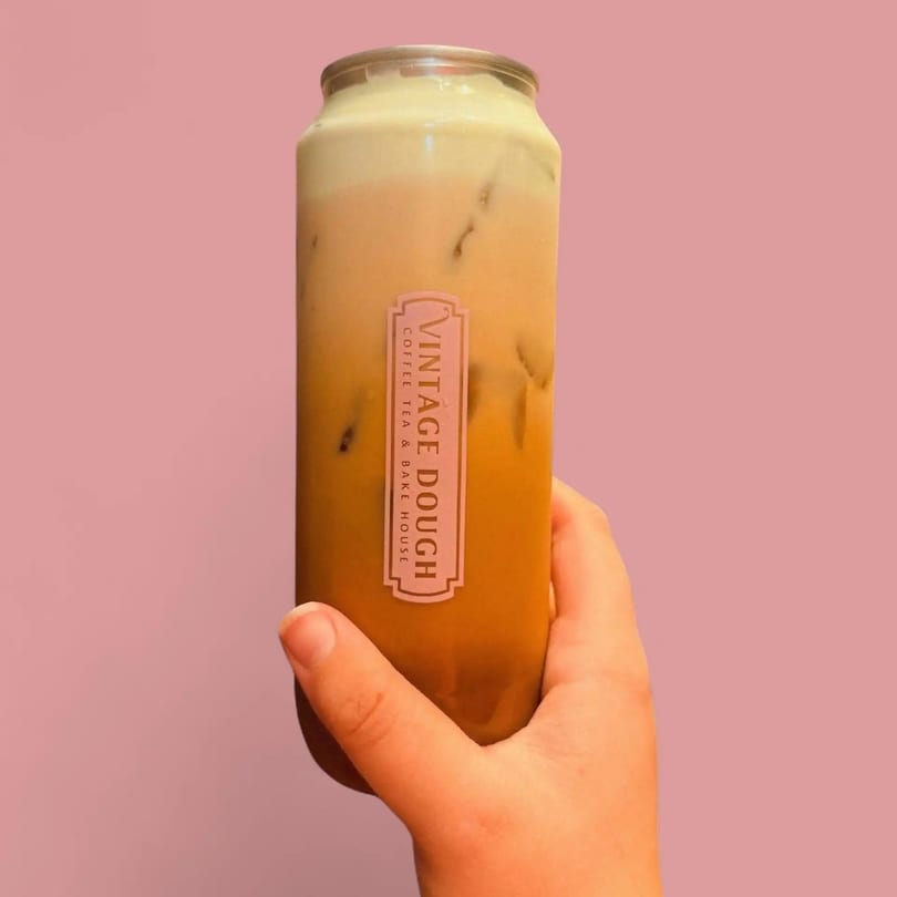Hand holding a Vintage Dough iced coffee can with a creamy top layer against a pink background.
