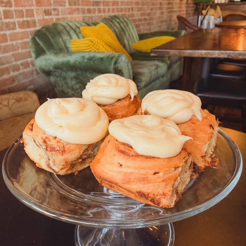Four cinnamon rolls with white icing on a glass cake stand in a cozy cafe setting.