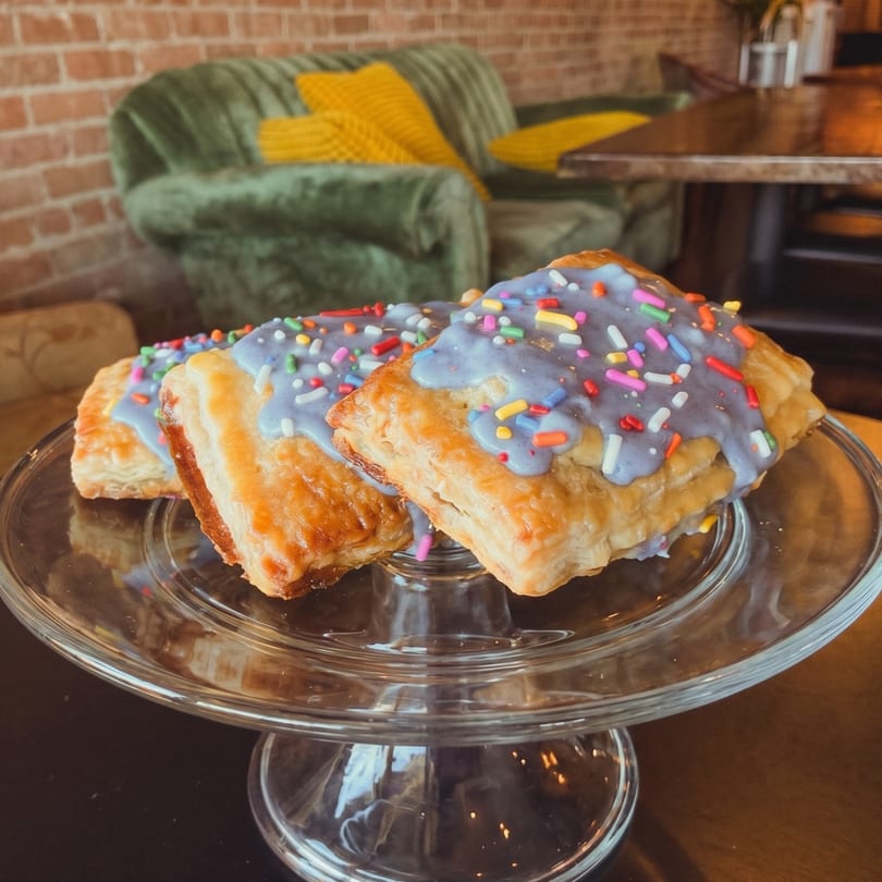 Three pastries with purple icing and colorful sprinkles on a glass cake stand in a cafe.